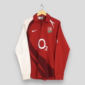 May include: A red and white long-sleeve rugby shirt with a collared neckline. The shirt features a white Nike swoosh, the O2 logo, and a floral emblem. The sleeves are white, and the body is red.