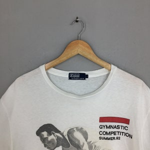 May include: White t-shirt featuring a graphic of a man performing gymnastics. The text "Gymnastic Competition Summer '92" is printed on the shirt. The shirt has a Polo by Ralph Lauren label.