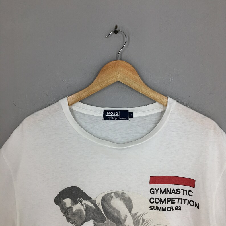 May include: White t-shirt featuring a graphic of a man performing gymnastics. The text "Gymnastic Competition Summer '92" is printed on the shirt. The shirt has a Polo by Ralph Lauren label.