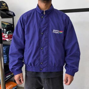 Puede incluir: Una chaqueta cortavientos de color morado con cuello alto, cierre de cremallera y tapeta frontal con botones a presión. En el pecho aparece el texto Benetton Formula 1 con una franja gráfica multicolor. La chaqueta tiene mangas largas y dobladillo elástico.