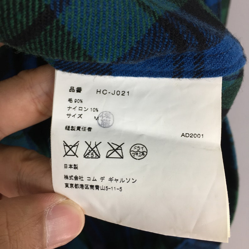 K&ouml;nnte beinhalten: Ein wei&szlig;es Etikett mit schwarzem Text und Pflegehinweisen. Das Etikett lautet "HC-J021", "Wolle 90%", "Nylon 10%", "Gr&ouml;&szlig;e M", "Hergestellt in Japan", "Comme des Gar&ccedil;ons", und "Tokio, Japan".