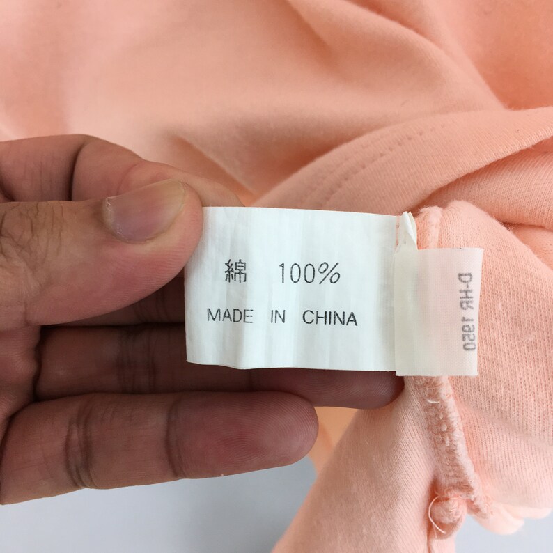 K&ouml;nnte beinhalten: Nahaufnahme eines pfirsichfarbenen Stoffes mit einem wei&szlig;en Etikett. Das Etikett zeigt "綿 100%" und "MADE IN CHINA" in schwarzer Schrift. Der Stoff wird von einer Hand gehalten.