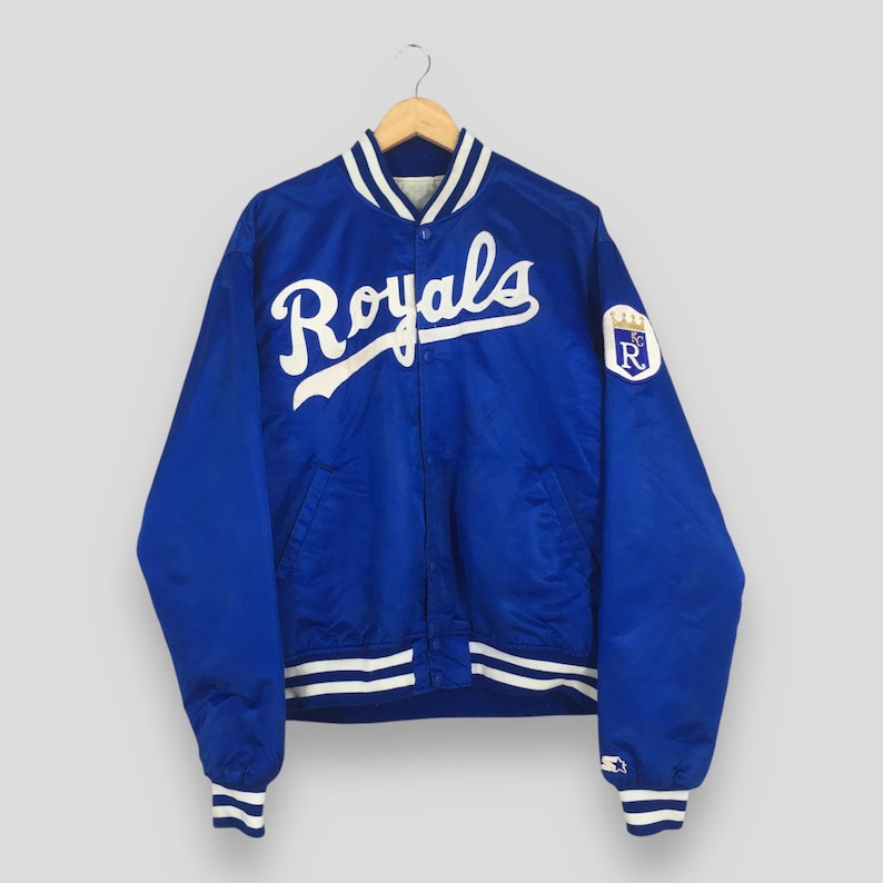 May include: A royal blue satin varsity jacket featuring the word Royals in white script across the chest. The jacket has a white and blue striped collar, cuffs, and hem, with a white and blue Royals logo patch on the left sleeve and snap buttons.