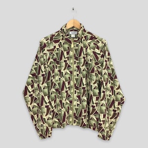 May include: A vintage zip-up jacket with a geometric pattern in shades of green, burgundy, and beige. The jacket has a high collar, long sleeves, and a gold-colored zipper. The jacket is hanging on a wooden hanger.