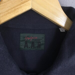 Puede incluir: Una camisa abotonada de color azul marino oscuro colgada de una percha de madera. La camisa tiene una etiqueta verde con texto rojo y blanco que dice "Yohji Yamamoto" y "A.A.R.". La camisa es talla "L".
