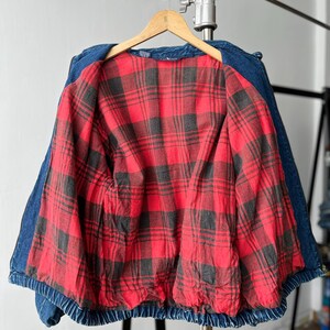 May include: A blue denim jacket featuring a vibrant red and black plaid flannel lining. The jacket is displayed on a wooden hanger and shows a classic collar, long sleeves, and a gathered elastic hem at the waist, providing a structured look.