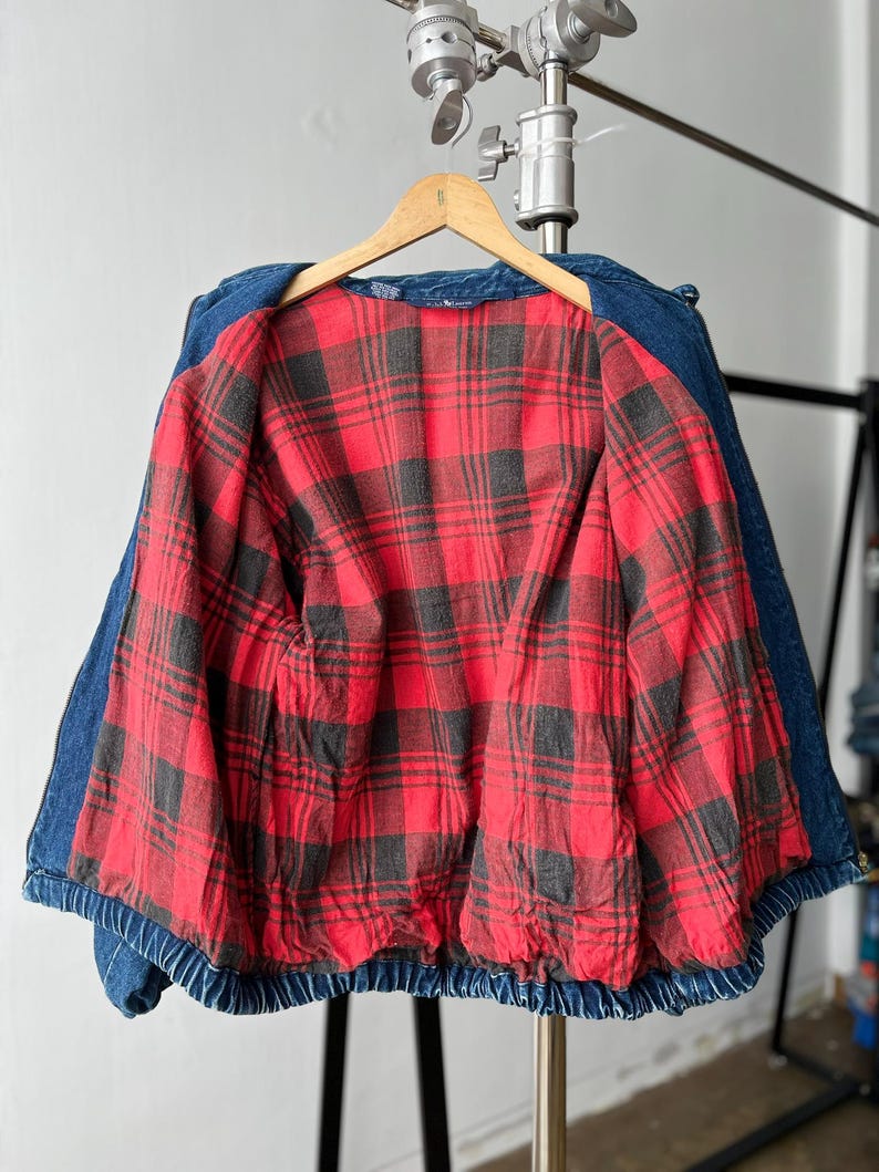 May include: A blue denim jacket featuring a vibrant red and black plaid flannel lining. The jacket is displayed on a wooden hanger and shows a classic collar, long sleeves, and a gathered elastic hem at the waist, providing a structured look.