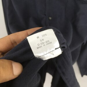 Puede incluir: Primer plano de una etiqueta de prenda azul marino. La etiqueta blanca tiene texto negro: "綿 100% SN C-OS-374 MADE IN JAPAN". La prenda es azul marino.