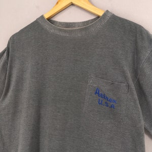 May include: A gray t-shirt with a pocket on the left chest. The pocket features the blue text "Avirex U.S.A." The shirt is hanging on a wooden hanger against a light gray background. The shirt has a crew neck and short sleeves.