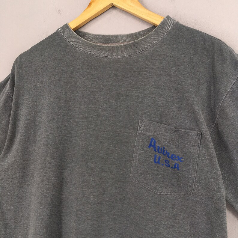 May include: A gray t-shirt with a pocket on the left chest. The pocket features the blue text "Avirex U.S.A." The shirt is hanging on a wooden hanger against a light gray background. The shirt has a crew neck and short sleeves.
