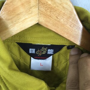 Pode incluir: Close-up de uma gola de camisa verde com uma etiqueta preta com caracteres japoneses e um emblema de drag&atilde;o. A etiqueta tamb&eacute;m inclui a palavra "GREATDRAGON" e o tamanho "L". A camisa est&aacute; pendurada em um cabide de madeira.