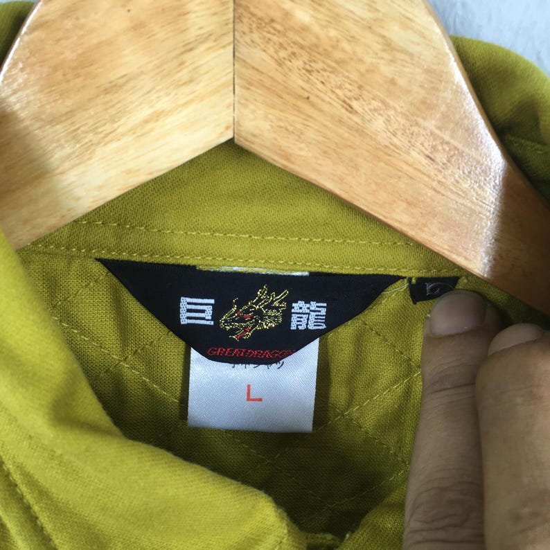 Pode incluir: Close-up de uma gola de camisa verde com uma etiqueta preta com caracteres japoneses e um emblema de drag&atilde;o. A etiqueta tamb&eacute;m inclui a palavra "GREATDRAGON" e o tamanho "L". A camisa est&aacute; pendurada em um cabide de madeira.