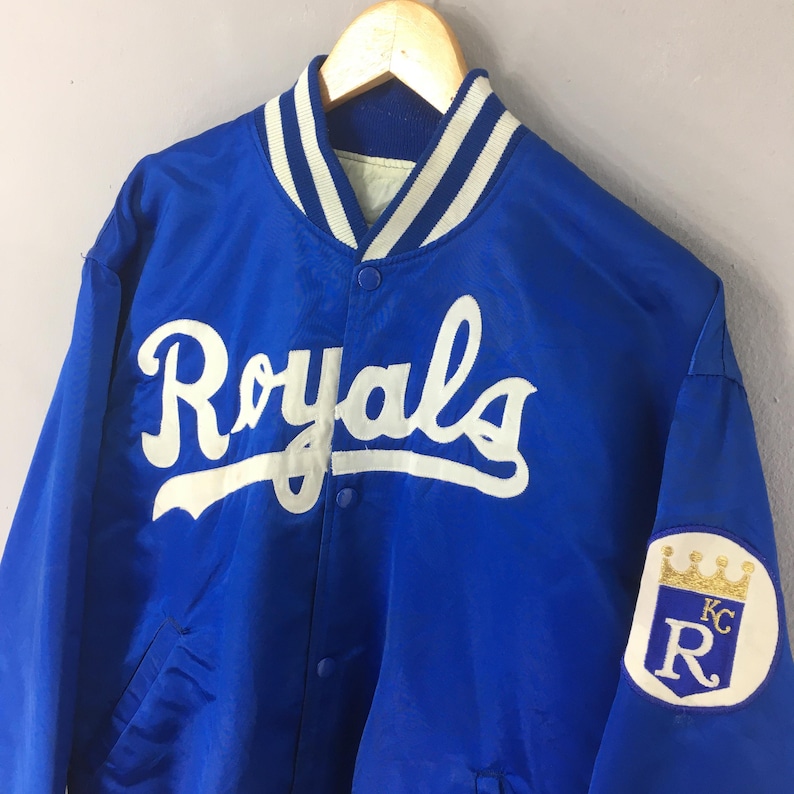May include: A royal blue satin varsity jacket featuring the word Royals in white script across the chest. The jacket has a ribbed collar with white stripes, snap button closures, and a white circular patch on the sleeve with a gold crown and KC R.