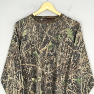 May include: Long-sleeved camouflage shirt with a crew neck. The shirt features a realistic forest pattern in shades of brown, green, and tan, with a tag on the collar. The shirt is hanging on a wooden hanger.