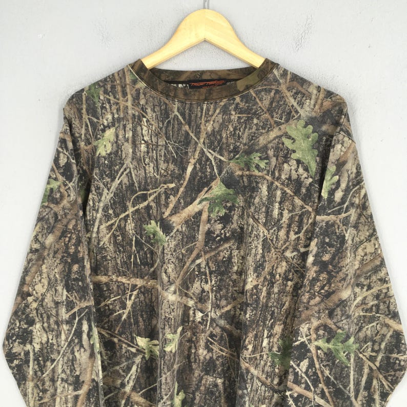 May include: Long-sleeved camouflage shirt with a crew neck. The shirt features a realistic forest pattern in shades of brown, green, and tan, with a tag on the collar. The shirt is hanging on a wooden hanger.