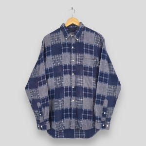 Puede incluir: Una camisa de manga larga con un diseño de patchwork en tonos azules y grises. La camisa tiene un cuello abotonado, puños abotonados y un bolsillo en el pecho. La tela parece ser un material suave y tejido, adecuado para ropa casual.