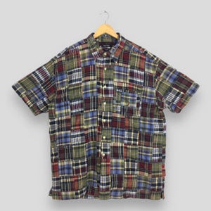 Puede incluir: Una camisa de manga corta con diseño de patchwork. La camisa presenta una variedad de patrones de cuadros en tonos de azul, verde, rojo y amarillo. La camisa está colgada de una percha de madera.