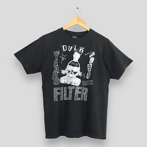 Puede incluir: Camiseta negra con un gráfico blanco que presenta una calavera, la palabra "Dulk", "Filter" y el texto "Ten Years Since 96". La camiseta está colgada en una percha de madera.