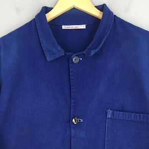 Peut inclure: Gros plan sur une veste de travail bleu cobalt avec une fermeture boutonn&eacute;e, un col et une seule poche. La veste est suspendue &agrave; un cintre en bois. Le tissu semble &ecirc;tre du coton robuste ou similaire.