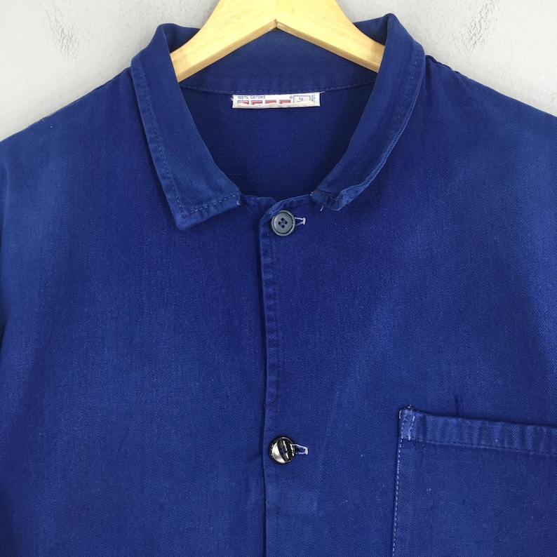 Peut inclure: Gros plan sur une veste de travail bleu cobalt avec une fermeture boutonn&eacute;e, un col et une seule poche. La veste est suspendue &agrave; un cintre en bois. Le tissu semble &ecirc;tre du coton robuste ou similaire.