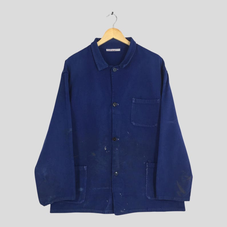 Peut inclure: Veste de travail bleu marine vintage avec une fermeture boutonn&eacute;e, un col et deux poches avant. La veste pr&eacute;sente des signes d'usure avec des &eacute;claboussures de peinture. Elle est suspendue &agrave; un cintre en bois.