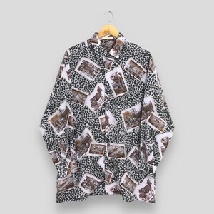 Könnte beinhalten: Ein langärmeliges Button-Down-Hemd mit einem einzigartigen Muster. Das Hemd hat einen weißen Hintergrund mit einem dunkelgrünen Fleckenmuster, überlagert mit rechteckigen Bildern von Tieren in Brauntönen. Das Hemd hängt an einem Holzbügel.
