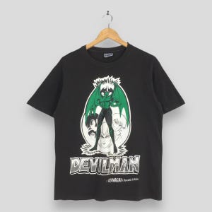 May include: Black t-shirt featuring a graphic of a muscular, winged figure with green wings and a white background. The word "DEVILMAN" is printed in a stylized font below the image, with text that reads "GO NAGAI & Dynamic Pro".