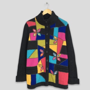 Puede incluir: Un cárdigan negro con un diseño de patchwork colorido. El cárdigan tiene cuello alto y cierre de botones. El diseño de patchwork presenta formas geométricas y bordados florales en tonos de rosa, azul, amarillo, verde y rojo.