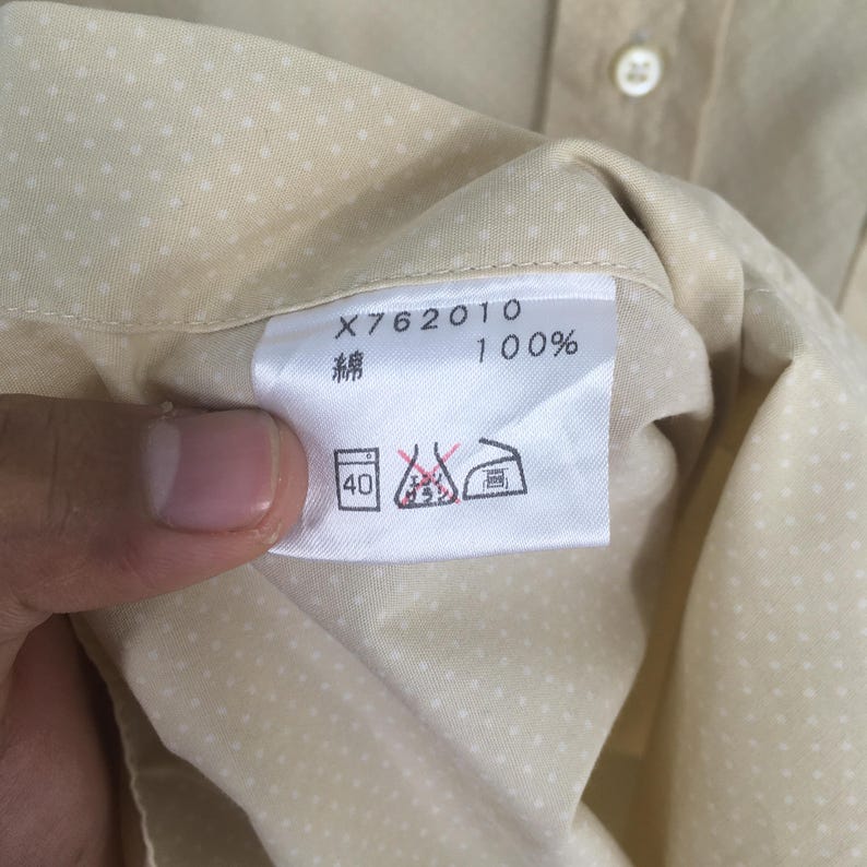 Pode incluir: Uma camisa bege com um padr&atilde;o de bolinhas brancas. A camisa tem uma etiqueta que diz "X762010 100% ". A etiqueta tamb&eacute;m tem um s&iacute;mbolo de instru&ccedil;&otilde;es de cuidados que indica que a camisa pode ser lavada na m&aacute;quina a 40 graus Celsius.