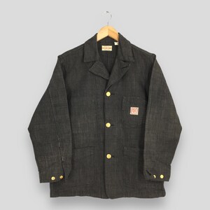Peut inclure: Veste en jean gris foncé avec un col classique et trois poches avant. La veste comporte des boutons dorés et un petit écusson avec du texte. La veste est suspendue à un cintre en bois.