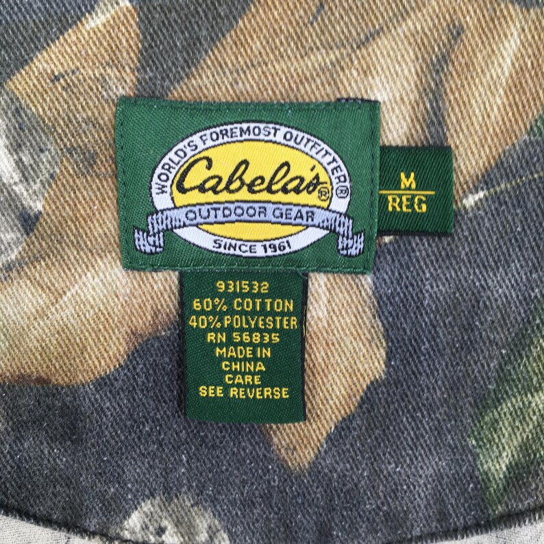 Op de afbeelding: Groen en bruin camouflagemateriaal met een groen label waarop "WORLD'S FOREMOST OUTFITTER Cabela's OUTDOOR GEAR SINCE 1961" staat en een kleiner groen label waarop "M REG" staat. Het label bevat ook de tekst "931532 60% COTTON 40% POLYESTER RN 56835 MADE IN CHINA CARE SEE REVERSE".
