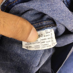 May include: Close-up of a blue denim fabric with a white tag. The tag displays care instructions, including "50" and "BLEACH". Other text includes "COTON COTTON" and "US RN 64961".