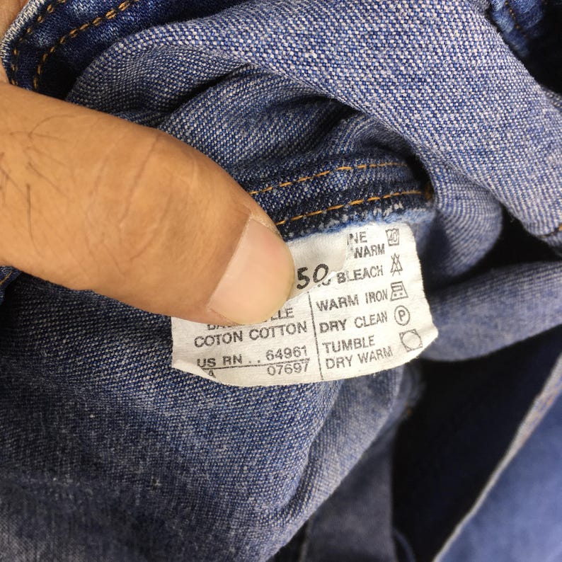 May include: Close-up of a blue denim fabric with a white tag. The tag displays care instructions, including "50" and "BLEACH". Other text includes "COTON COTTON" and "US RN 64961".