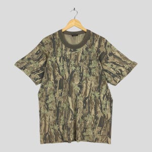 以下が含まれることがあります： 茶色、緑、ベージュの色合いのカモフラージュ柄の半袖Tシャツ。クルーネックでゆったりとしたフィット感。葉のモチーフを取り入れ、自然でアウトドアな雰囲気を演出しています。