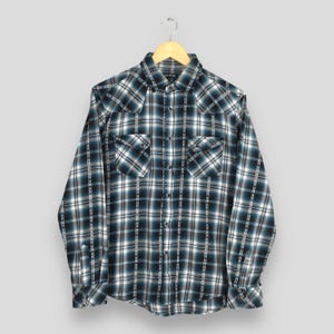 Puede incluir: Una camisa de manga larga a cuadros en tonos de verde azulado, negro y blanco, colgada de una percha de madera. La camisa tiene dos bolsillos en el pecho y puños abotonados. El patrón de cuadros es un diseño clásico.