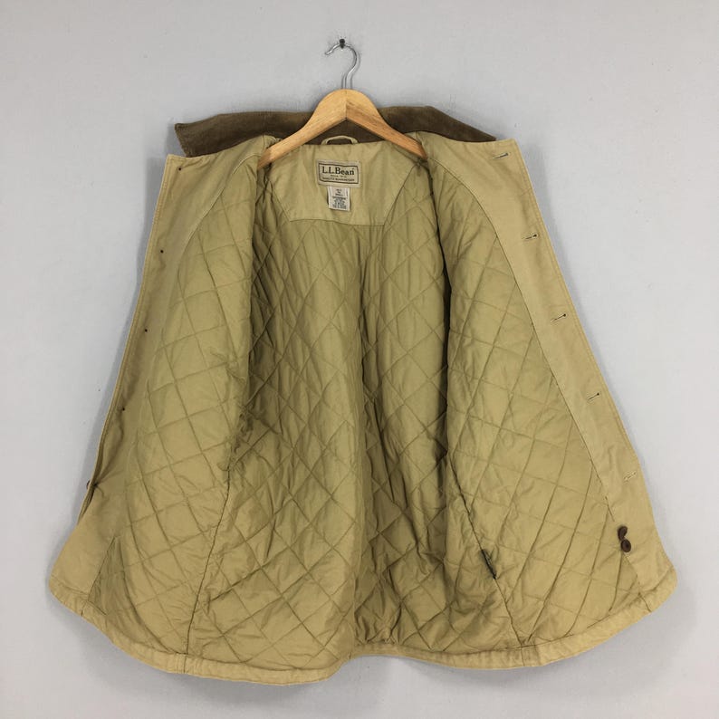 May include: Tan quilted jacket with a brown corduroy collar, hanging on a wooden hanger. The jacket is open, revealing a diamond-patterned interior lining. The label reads "L.L.Bean".