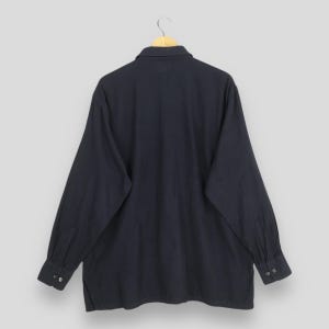 Puede incluir: Una camisa de manga larga azul marino con cuello y pu&ntilde;os abotonados. La camisa est&aacute; colgada en una percha de madera sobre un fondo gris claro. Se ve la parte trasera de la camisa, mostrando los detalles de los botones en los pu&ntilde;os.