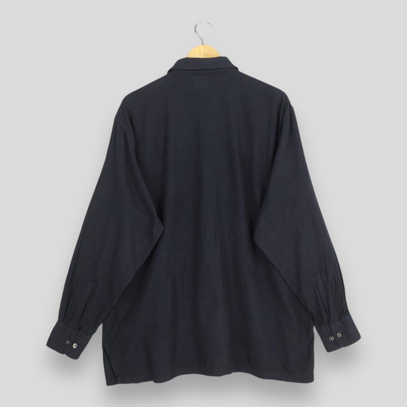 Puede incluir: Una camisa de manga larga azul marino con cuello y pu&ntilde;os abotonados. La camisa est&aacute; colgada en una percha de madera sobre un fondo gris claro. Se ve la parte trasera de la camisa, mostrando los detalles de los botones en los pu&ntilde;os.