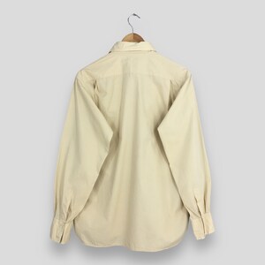 Pode incluir: Uma camisa de manga comprida bege claro com colarinho e bot&otilde;es. A camisa est&aacute; pendurada em um cabide de madeira.