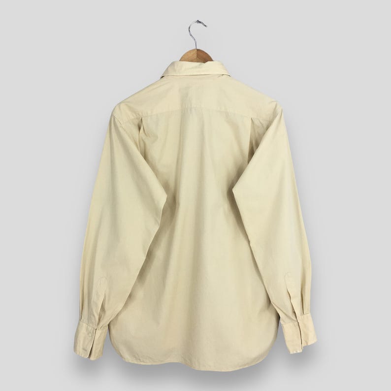 Pode incluir: Uma camisa de manga comprida bege claro com colarinho e bot&otilde;es. A camisa est&aacute; pendurada em um cabide de madeira.