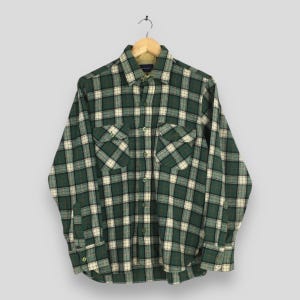 May include: A green and white checked shirt with long sleeves and two chest pockets, hanging on a wooden hanger. The shirt has a button-down front and a classic collar. The pattern is a grid of green and white squares.