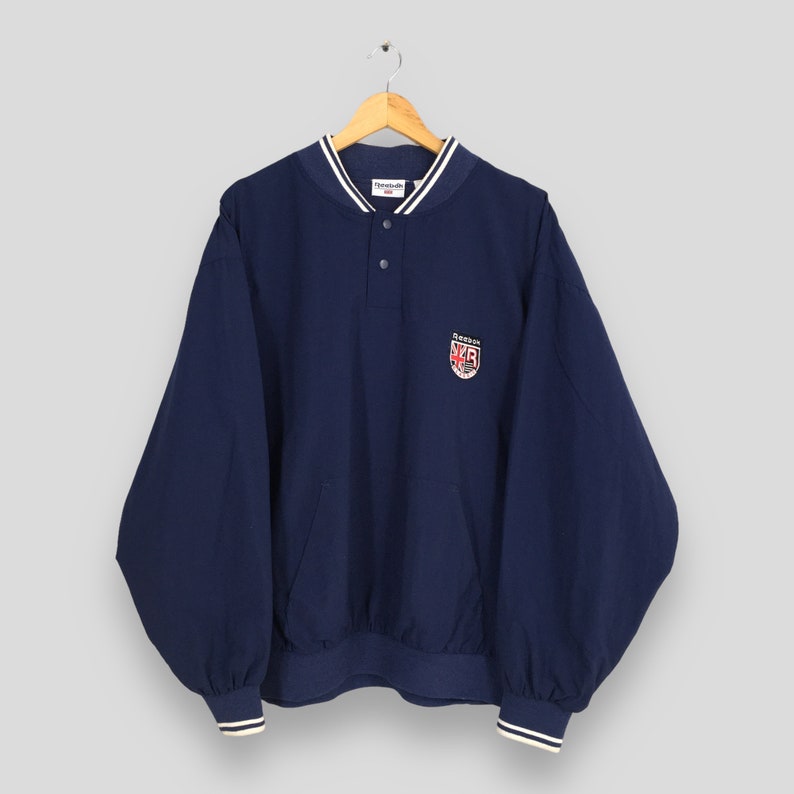 May include: A navy blue Reebok windbreaker with a white and navy blue striped collar and cuffs. The windbreaker has a button closure and a small embroidered logo on the left chest that says "Reebok" and "425".