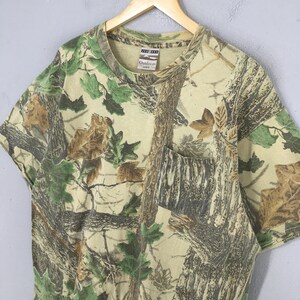 May include: A short-sleeved camouflage t-shirt with a chest pocket. The shirt features a pattern of brown tree trunks and green and brown leaves. The tag reads "Outdoors".