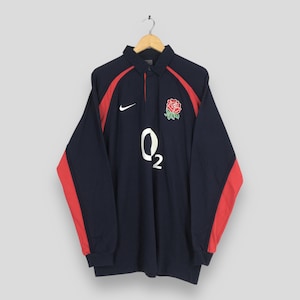 Op de afbeelding: Marineblauw rugby shirt met lange mouwen en rode accenten op de mouwen en schouders. Voorzien van een witte "O2"-afbeelding en een rood rozenembleem. Inclusief een Nike-logo. Het shirt hangt aan een houten hanger.