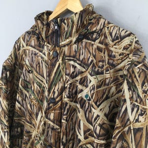 Puede incluir: Chaqueta de camuflaje con cuello alto, cremallera y cierre de botones. La chaqueta presenta un patr&oacute;n de color marr&oacute;n, beige y verde que se asemeja a juncos. Un peque&ntilde;o logotipo verde y blanco es visible en la parte delantera.