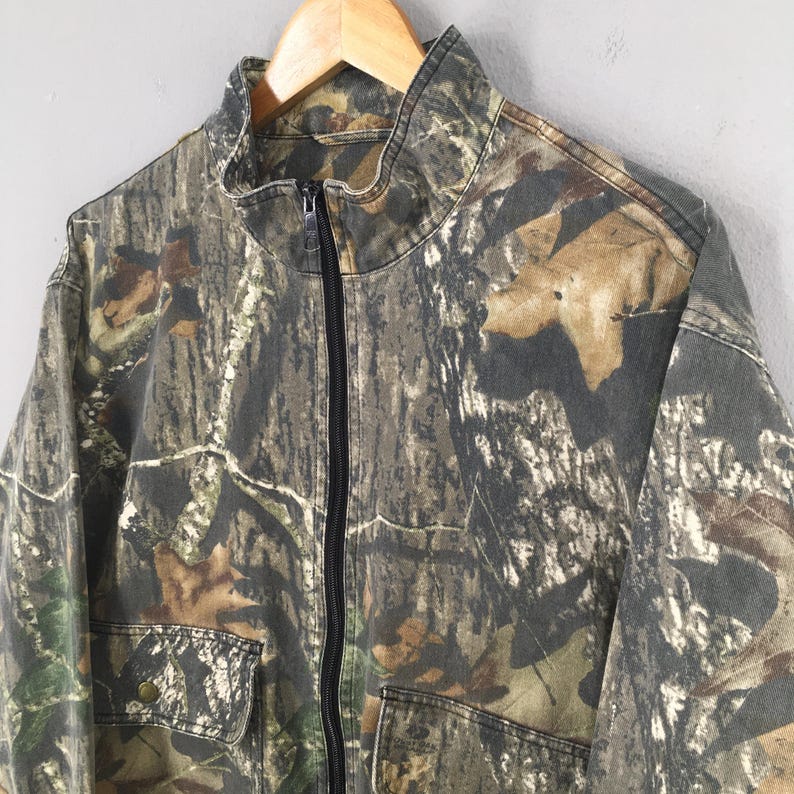 Op de afbeelding: Een camouflage-printjas met een ritssluiting. De jas heeft een opstaande kraag en twee grote zakken.