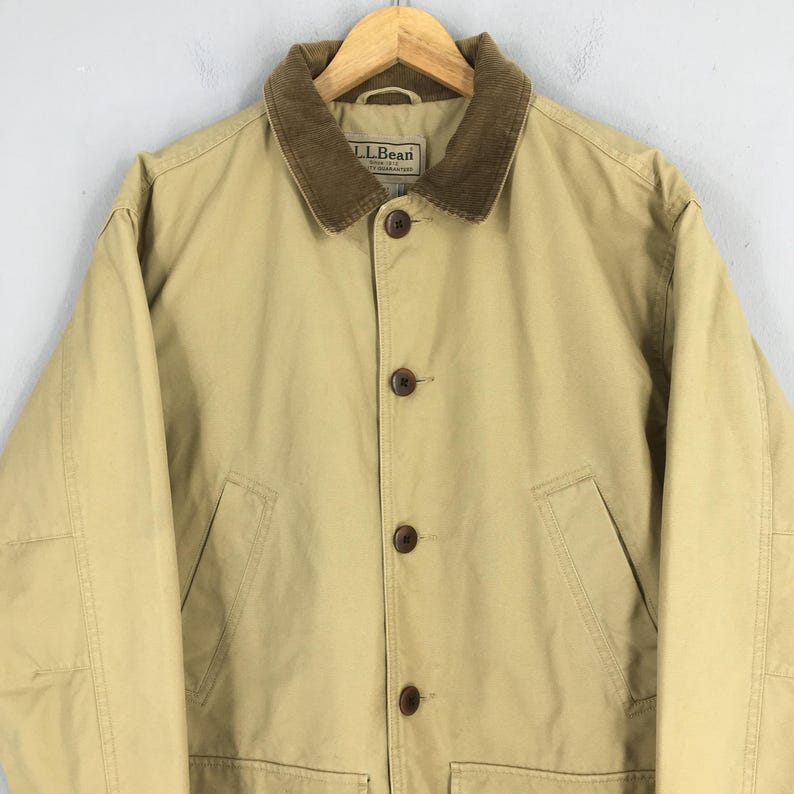 May include: Tan jacket with a brown corduroy collar and button closure. The jacket has a label that reads "L.L.Bean". The jacket has two front pockets and a classic design.
