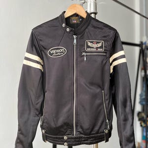 Könnte beinhalten: Eine schwarze Jacke mit Stehkragen, durchgehendem Frontreißverschluss und zwei cremefarbenen Streifen an jedem Ärmel. Auf der Brust befinden sich ein Vanson Leathers-Aufnäher und ein American Bass-Aufnäher mit rotem Stern. Mit Reißverschlusstaschen.