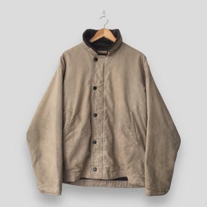 May include: A tan canvas jacket featuring a dark brown faux fur lining, a high collar, and a front placket with four black buttons. The jacket has long sleeves, two side pockets, and a structured fit, displayed on a wooden hanger.