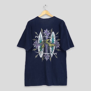 May include: A navy blue t-shirt with a graphic print of a cross with two surfboards on either side. The graphic is in shades of green, purple, and gold.